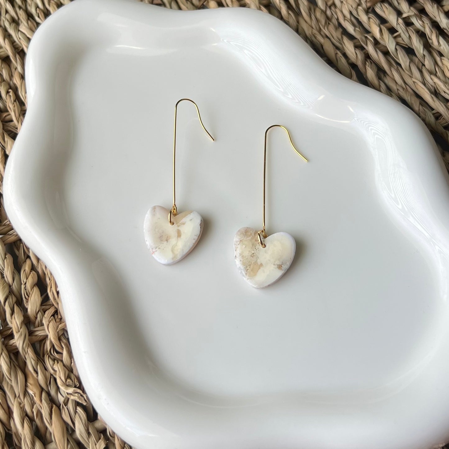 Heart-shaped clay earrings with elongated gold stainless steel wires on a white dish with a woven background