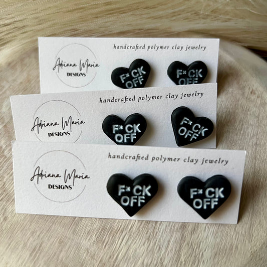 Handmade polymer clay stud earrings with black hearts and white text, displayed on branded white cards. Text on heart clay earrings are F*CK OFF