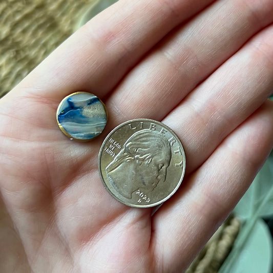 Hand holding a polymer clay circle stud earring in ocean blue with gold edges, lightweight and coastal-inspired and quarter to show size comparison.