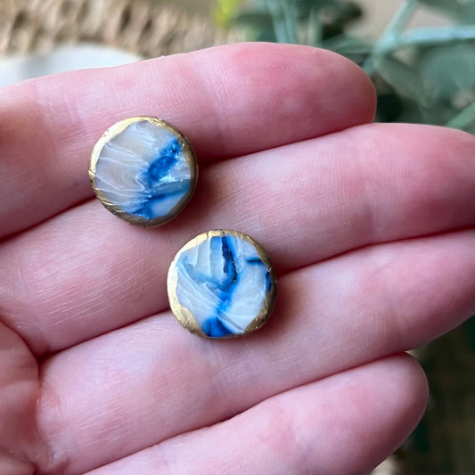Two marbled blue and white polymer clay circle stud earrings with gold accents held between fingers against a blurred natural background.
