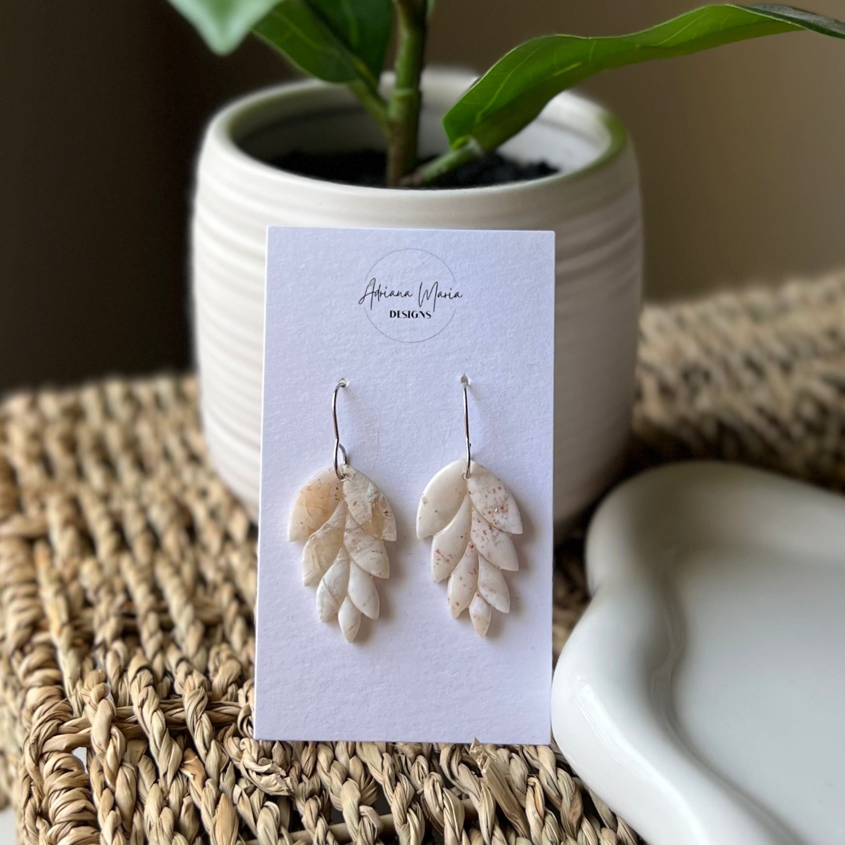 Pair of leaf-shaped earrings on a branded card with a plant and ceramic dish in the background.