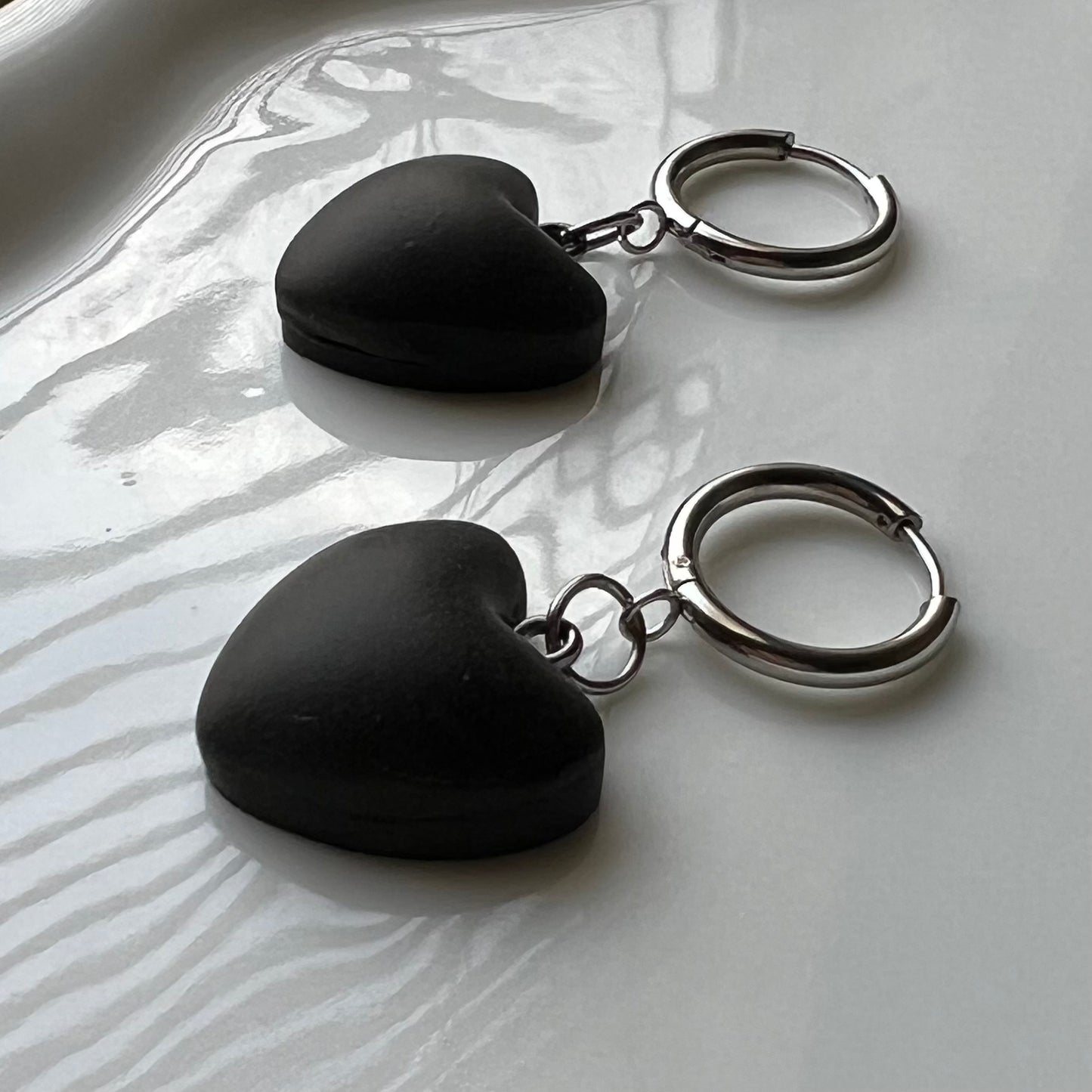 Black heart-shaped clay earrings with circle huggie hoops on a white dish with a natural background