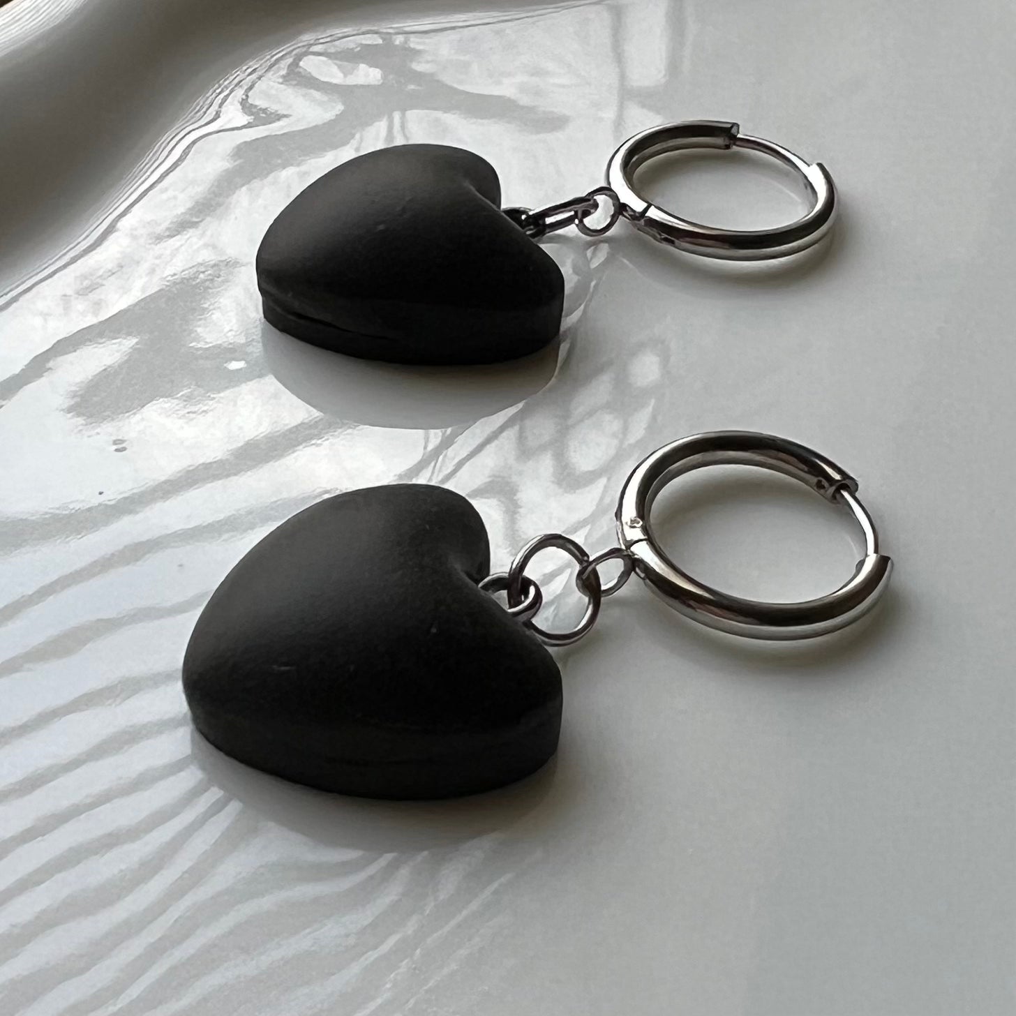 Black heart-shaped clay earrings with circle huggie hoops on a white dish with a natural background