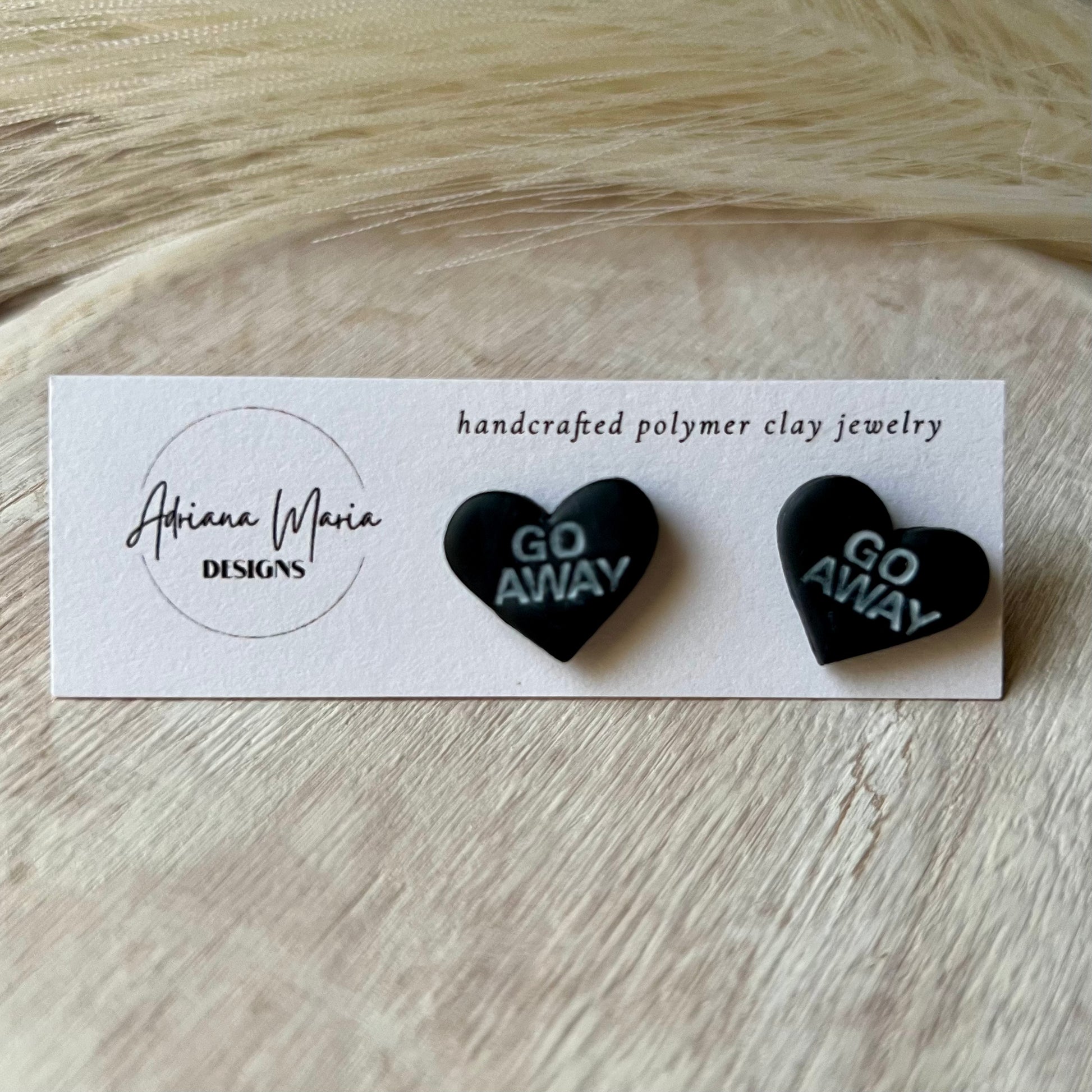 Handmade polymer clay stud earrings with black hearts and white text, displayed on branded white cards. Text on heart clay earrings is GO AWAY