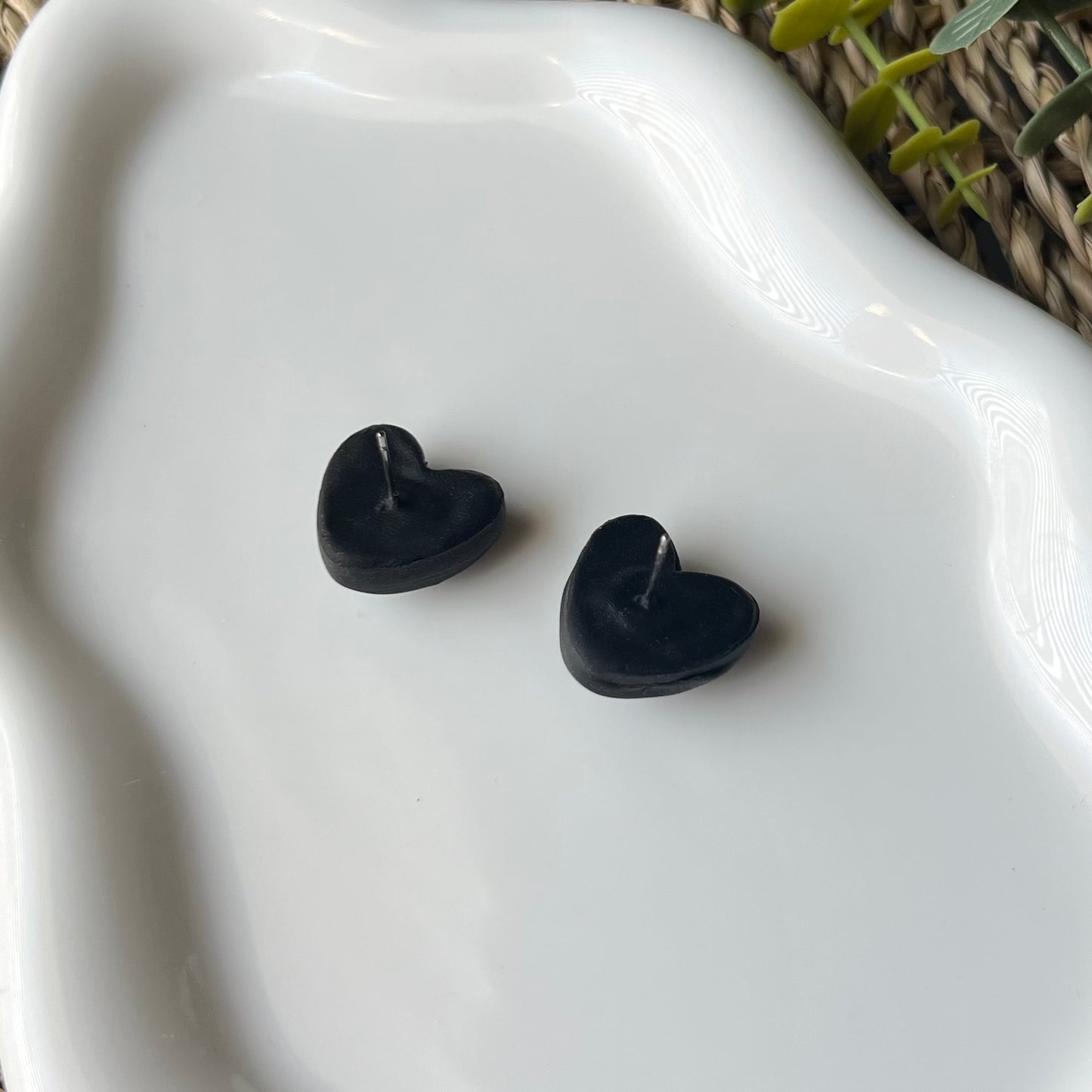 Black heart-shaped earrings showing stainless steel posts embedded in the back with clay on a white dish with a natural background