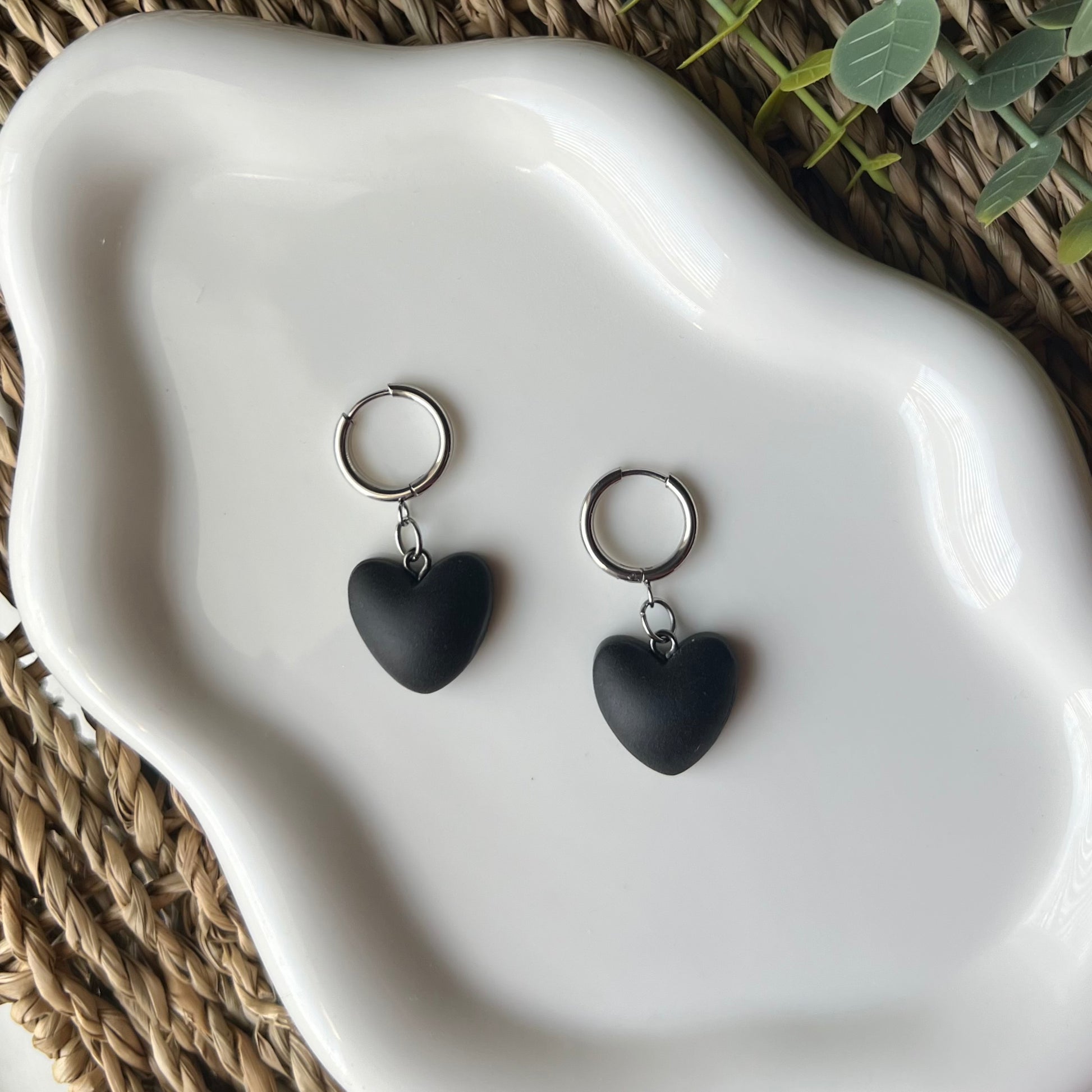 Black heart-shaped clay earrings with circle huggie hoops on a white dish with a natural background